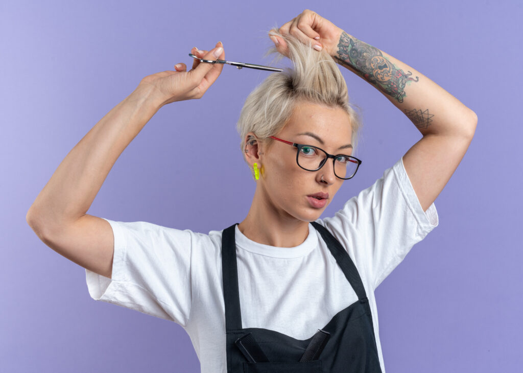 mulher jovem e bonita com um corte de cabelos curtos, com coloração loira, segurando uma tesoura em uma das mãos simulando seu próprio corte de cabelo.