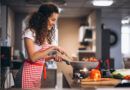 Jovem do sexo feminino com cabelos encaracolados, usando avental xadrez de vermelho, feliz com panela cozinhando. Remetendo ao tema do artigo: Segredos da Cozinha