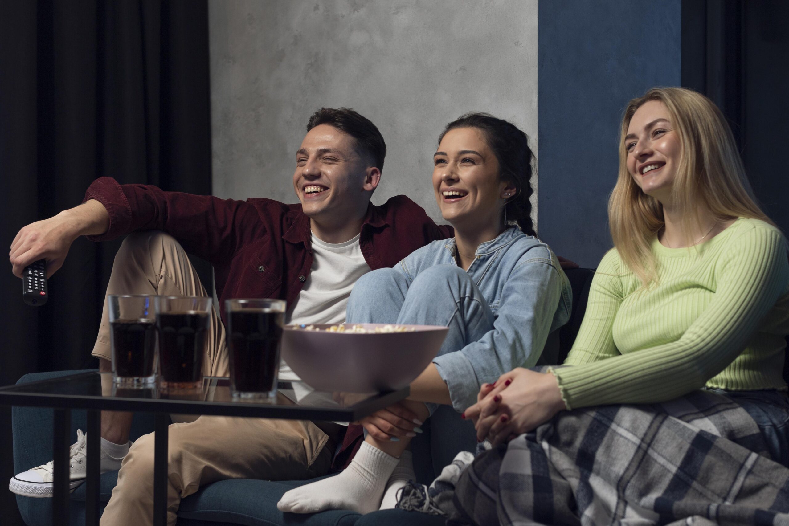 duas jovens com um rapaz  todos sorrindo sentados em um sofá com bacia de pipoca, e refrigerantes de cola assistindo tv
