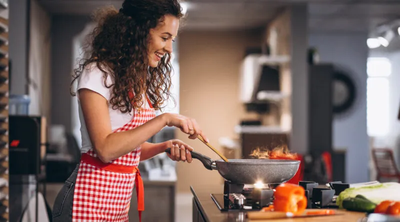 Jovem do sexo feminino com cabelos encaracolados, usando avental xadrez de vermelho, feliz com panela cozinhando. Remetendo ao tema do artigo: Segredos da Cozinha